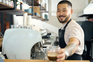 Handsome young barista smiling at the camera while offering a cup of fresh coffee to taste